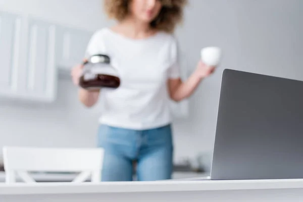 Partial view of blurred woman with cup and coffee pot near laptop in kitchen — Stock Photo