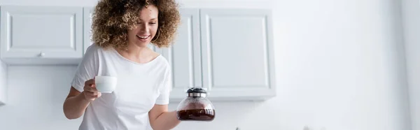 Mujer feliz con el pelo ondulado taza de celebración y cafetera en la cocina, pancarta — Stock Photo