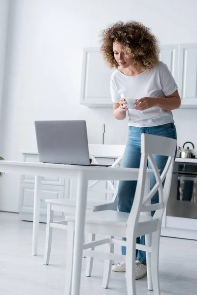 Mulher encaracolado com xícara de café em pé perto de laptop na cozinha — Fotografia de Stock