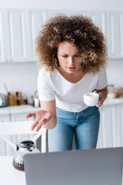 Femme bouclée avec tasse de café pointant avec la main pendant l'appel vidéo sur ordinateur portable dans la cuisine — Photo de stock