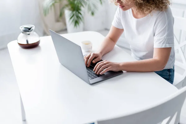 Vista parcial de la mujer en camiseta blanca escribiendo en el ordenador portátil cerca de taza y cafetera - foto de stock