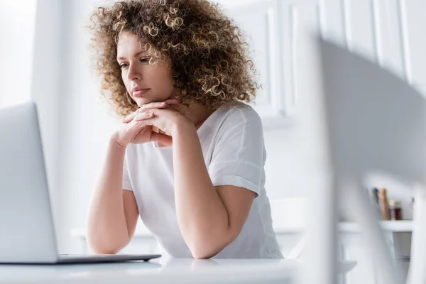 Mujer reflexiva con las manos apretadas sentado cerca de la computadora portátil en primer plano borrosa - foto de stock