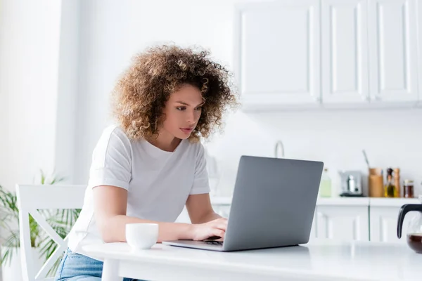 Femme concentrée avec cheveux bouclés tapant sur ordinateur portable près tasse de café dans la cuisine — Photo de stock