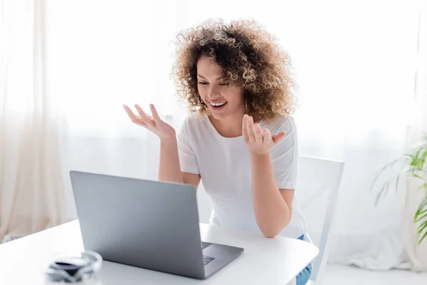 Fröhliche Frau mit welligem Haar gestikuliert beim Videochat am heimischen Laptop — Stockfoto