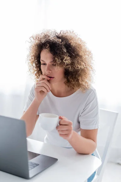 Mulher pensativa com xícara de café pensando perto de laptop na cozinha — Fotografia de Stock