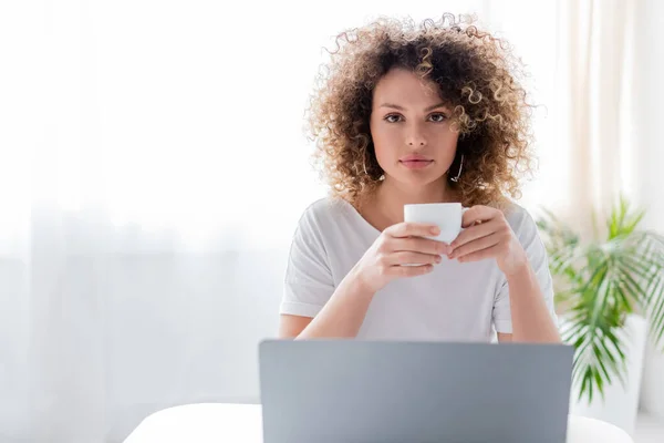 Pretty and curly woman holding coffee cup and looking at camera near laptop — Stock Photo