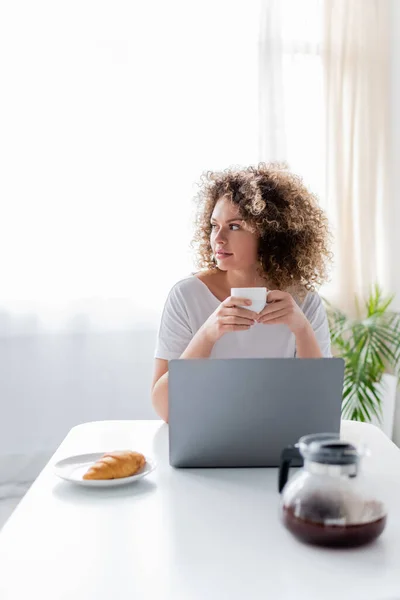 Donna riccia seduta con tazza di caffè vicino al computer portatile e gustoso croissant in cucina — Foto stock