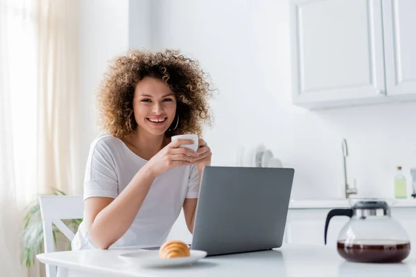 Mujer alegre con taza de café mirando a la cámara cerca del ordenador portátil y croissant — Stock Photo