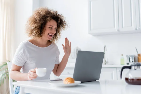 Donna felice con tazza di caffè mano agitando vicino al computer portatile e croissant sfocato — Foto stock