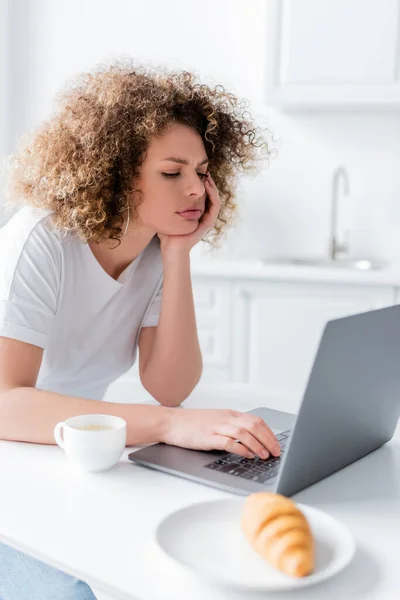 Mujer pensativa con la mano cerca de la cara utilizando el ordenador portátil cerca de la taza de café - foto de stock