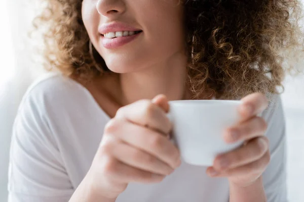 Частичный вид улыбающейся женщины с кудрявыми волосами, держащей чашку белого кофе — стоковое фото