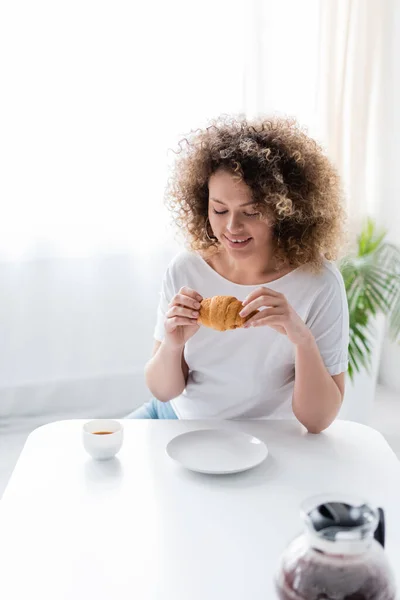 Mulher sorridente com delicioso croissant perto da xícara e panela de café burred — Fotografia de Stock