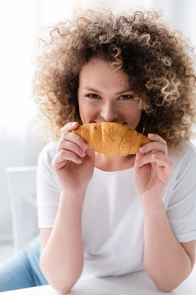 Mulher satisfeita com cabelo ondulado cheirando delicioso croissant e olhando para a câmera — Fotografia de Stock