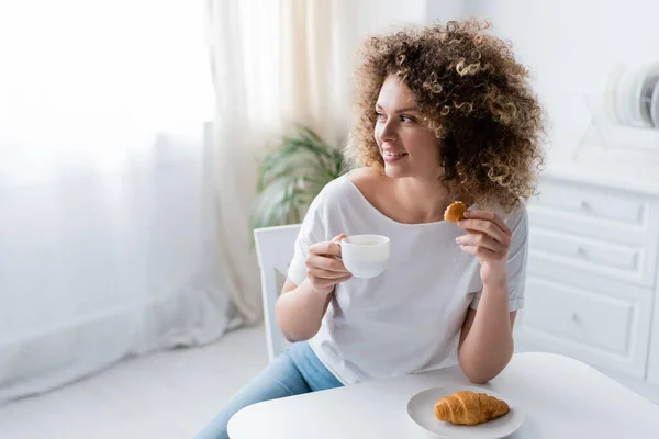 Happy woman with croissant and cup of coffee looking away in kitchen — Stock Photo