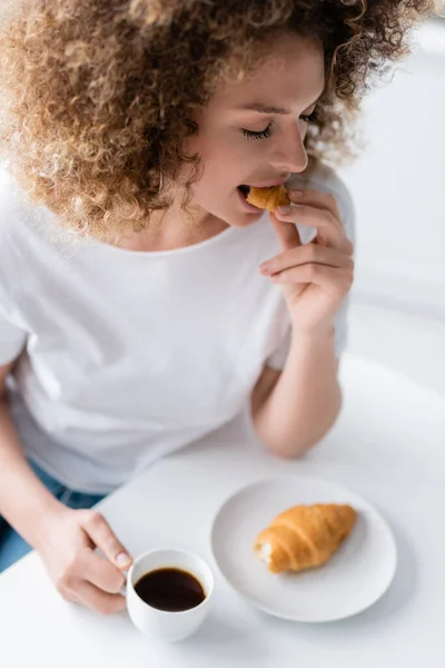 Mulher encaracolado com olhos fechados comer croissant enquanto segurando xícara de café — Fotografia de Stock