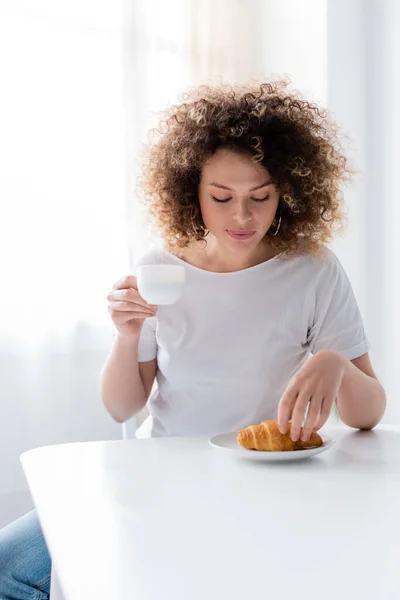 Mujer rizada en camiseta blanca sosteniendo taza de café cerca de sabroso croissant - foto de stock