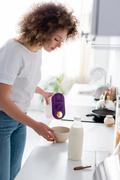 Curly woman holding container with corn flakes near bowl and bottle of milk — Stock Photo
