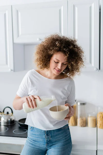 Mujer sonriente con el pelo ondulado verter leche fresca de la botella en el tazón en la cocina - foto de stock