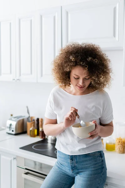 Donna felice con i capelli ricci tenendo cucchiaio e ciotola di fiocchi di mais gustosi in cucina — Foto stock