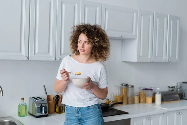 Mujer rizada en camiseta blanca sosteniendo tazón y cuchara con hojuelas de maíz en la cocina — Stock Photo