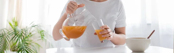 Cropped view of woman pouring orange juice from jug into glass near bowl, banner — Stock Photo