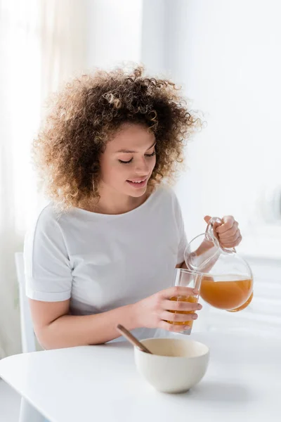 Mujer joven y positiva con el pelo ondulado vertiendo jugo de naranja de la jarra en el vidrio - foto de stock