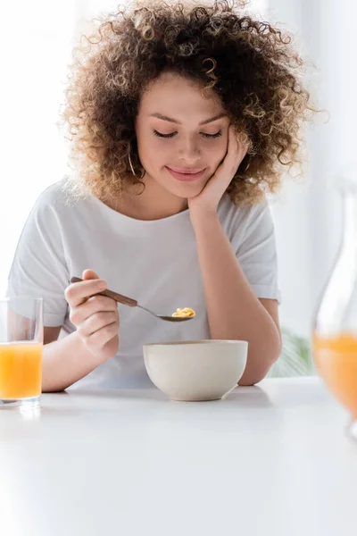 Задоволена кучерява жінка їсть кукурудзяні пластівці біля склянки свіжого апельсинового соку — стокове фото