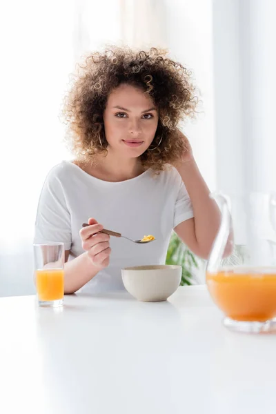 Donna positiva con capelli ondulati che tengono cucchiaio di fiocchi di mais vicino al succo d'arancia fresco — Foto stock