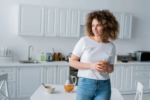 Mulher encaracolado com vidro de suco de laranja fresco sorrindo na cozinha turva — Fotografia de Stock