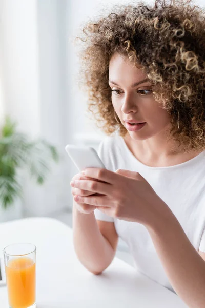 Femme stupéfaite avec la messagerie ondulée de cheveux sur le téléphone mobile près du verre de jus d'orange — Photo de stock