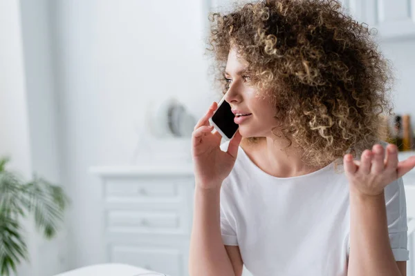 Femme bouclée en blanc t-shirt geste tout en parlant sur smartphone à la maison — Photo de stock