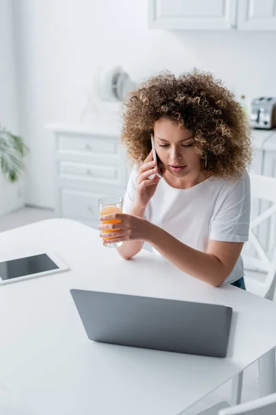 Mulher encaracolado com vidro de suco de laranja falando no celular perto do computador na cozinha — Fotografia de Stock