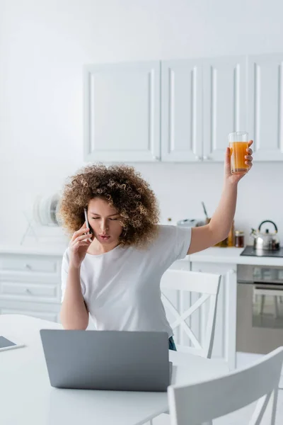 Donna eccitata brindare con vetro di succo d'arancia mentre parla su smartphone vicino al computer portatile in cucina — Foto stock
