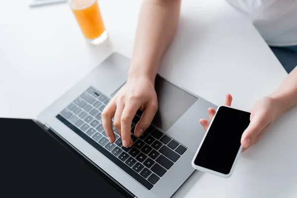 Vista parcial de la mujer con el teléfono móvil escribiendo en el ordenador portátil cerca del vaso de jugo de naranja - foto de stock