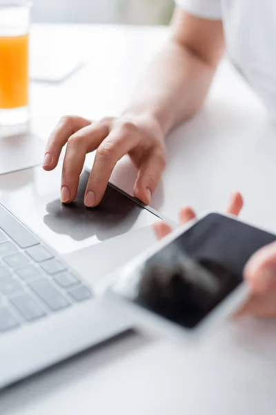 Partial view of woman using laptop and holding blurred smartphone with blank screen — Stock Photo