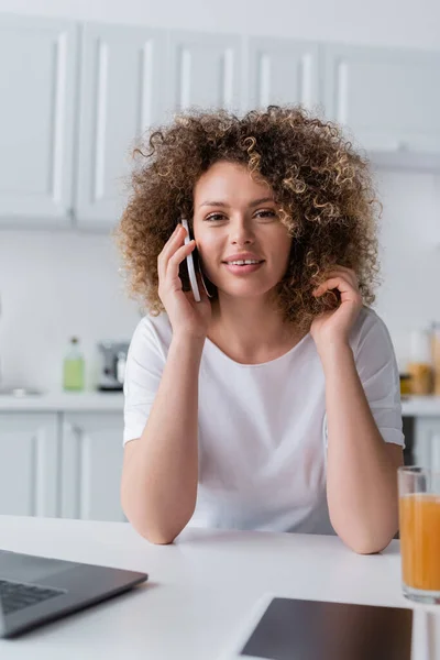 Mujer rizada feliz hablando en el teléfono móvil cerca de la tableta digital y el vaso de jugo de naranja - foto de stock