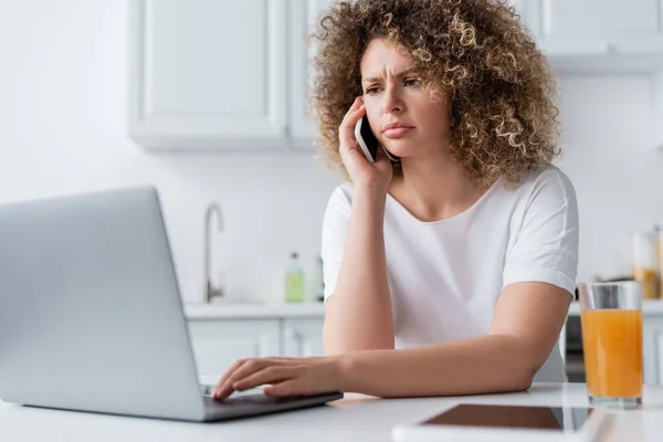 Mujer seria y frunciendo el ceño usando el ordenador portátil y hablando por teléfono celular en la cocina - foto de stock