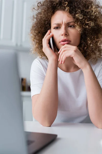 Serious woman frowning while talking on smartphone near blurred laptop — Stock Photo