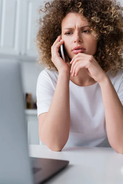 Mujer seria frunciendo el ceño mientras habla en el teléfono inteligente cerca de la computadora portátil borrosa - foto de stock