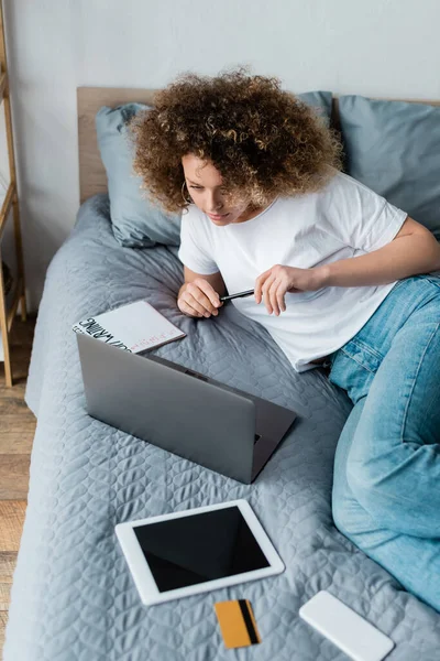 Jovem copywriter olhando para laptop perto de tablet digital e notebook na cama — Fotografia de Stock