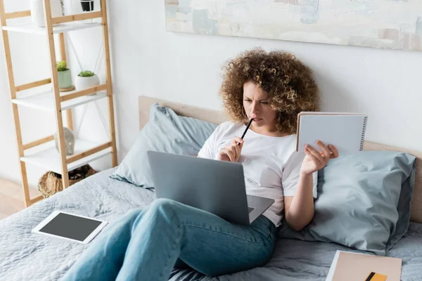 Nachdenkliche Frau mit Stift und Notizbuch arbeitet auf dem Bett in der Nähe des digitalen Tablets — Stockfoto