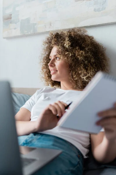 Mulher encaracolado sorridente com notebook olhando para longe perto laptop borrado no quarto — Fotografia de Stock