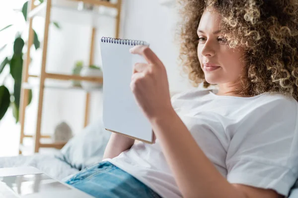 Positive Frau mit lockigem Haar schaut zu Hause auf Notizbuch im Bett — Stockfoto