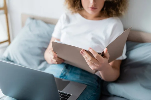 Partial view of woman with folder and laptop sitting on bed on blurred background — Stock Photo