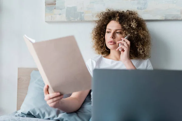 Jeune femme bouclée avec dossier parlant sur smartphone près d'un ordinateur portable dans la chambre — Photo de stock