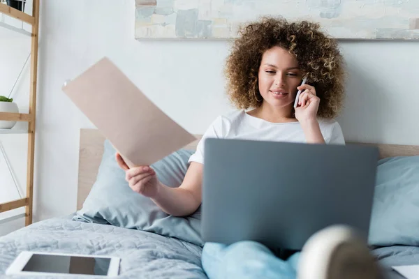 Mujer rizada con carpeta que habla en el teléfono inteligente cerca del ordenador portátil y la tableta digital en la cama - foto de stock