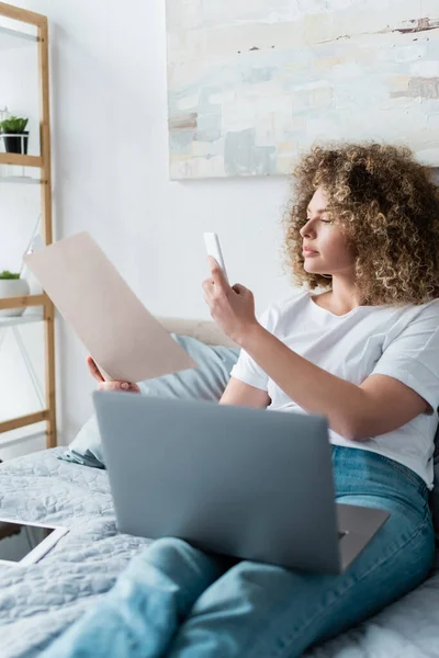 Mujer rizada tomando fotos de documentos en el teléfono inteligente cerca de la computadora portátil en el dormitorio - foto de stock