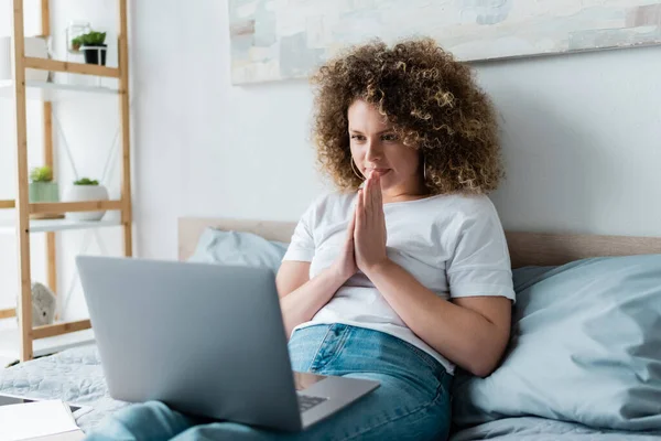 Positive woman sitting with praying hands near laptop in bedroom — Stock Photo
