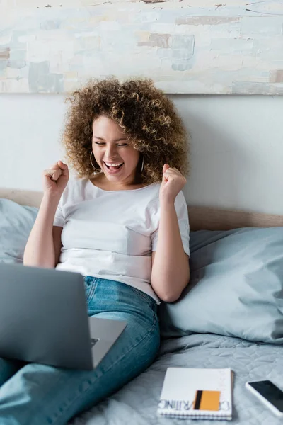 Excited woman showing success gesture near laptop while sitting on bed at home — Stock Photo