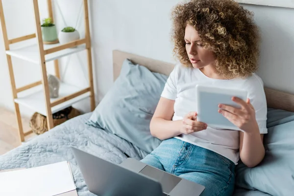 Serious woman with digital tablet looking at laptop while sitting on bed — Stock Photo